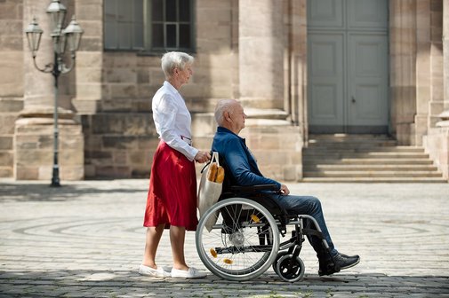 Frau schiebt ihren Mann Eine Seniorin schiebt ihren Mann, der im Rollstuhl sitzt, über einen Platz