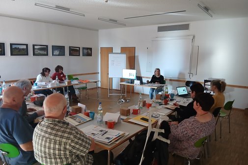 in einem Raum sitzen viele Menschen an Tischen und hören einer Referentin zu