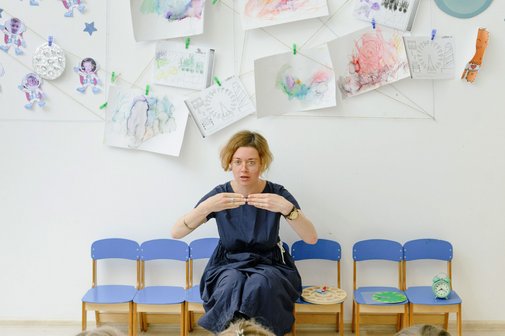 drei Kinder sitzen auf Stühlen, gegenüber eine Lehrerin